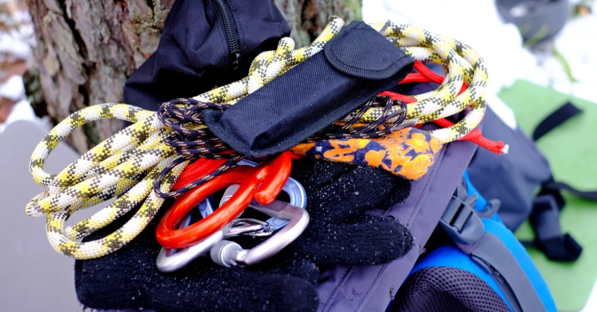 A close-up of some rope, carabiners, gloves, and other items piled on top of a backpack resting against a tree.