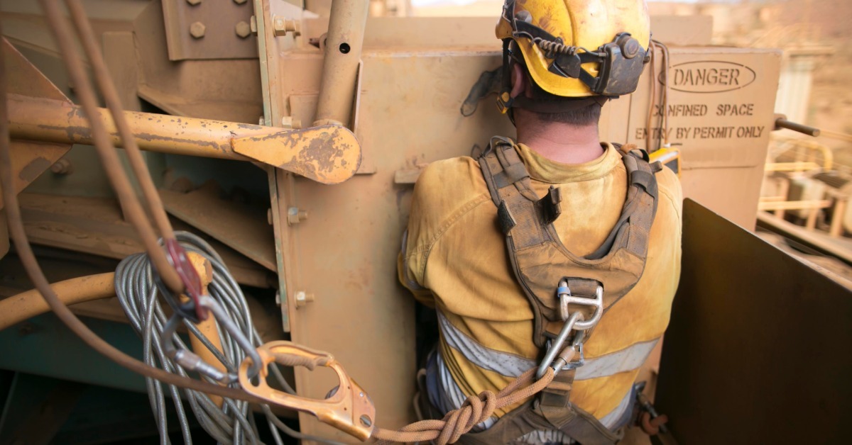 A close-up of a worker wearing a yellow helmet and a safety harness connected with ropes to industrial machinery.