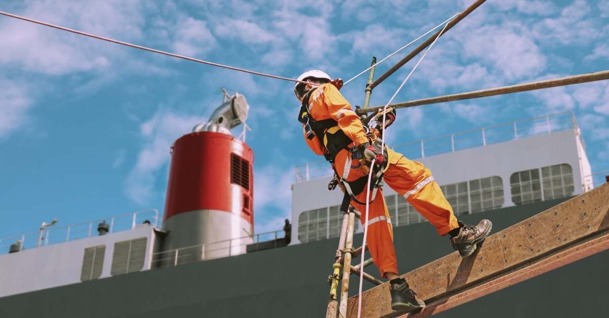 A worker in orange safety gear secured by ropes and a harness while working at height beside a large structure.
