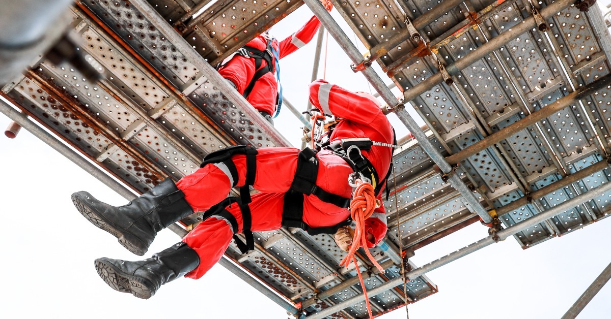 A worker in red protective gear and a safety harness suspended by ropes beneath elevated metal scaffolding.