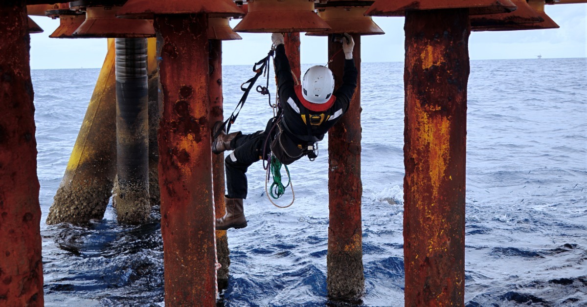 A worker wearing a white hard hat and safety harness while suspended between metal support columns above ocean water. A worker wearing a white hard hat and safety harness while suspended between metal support columns above ocean water.