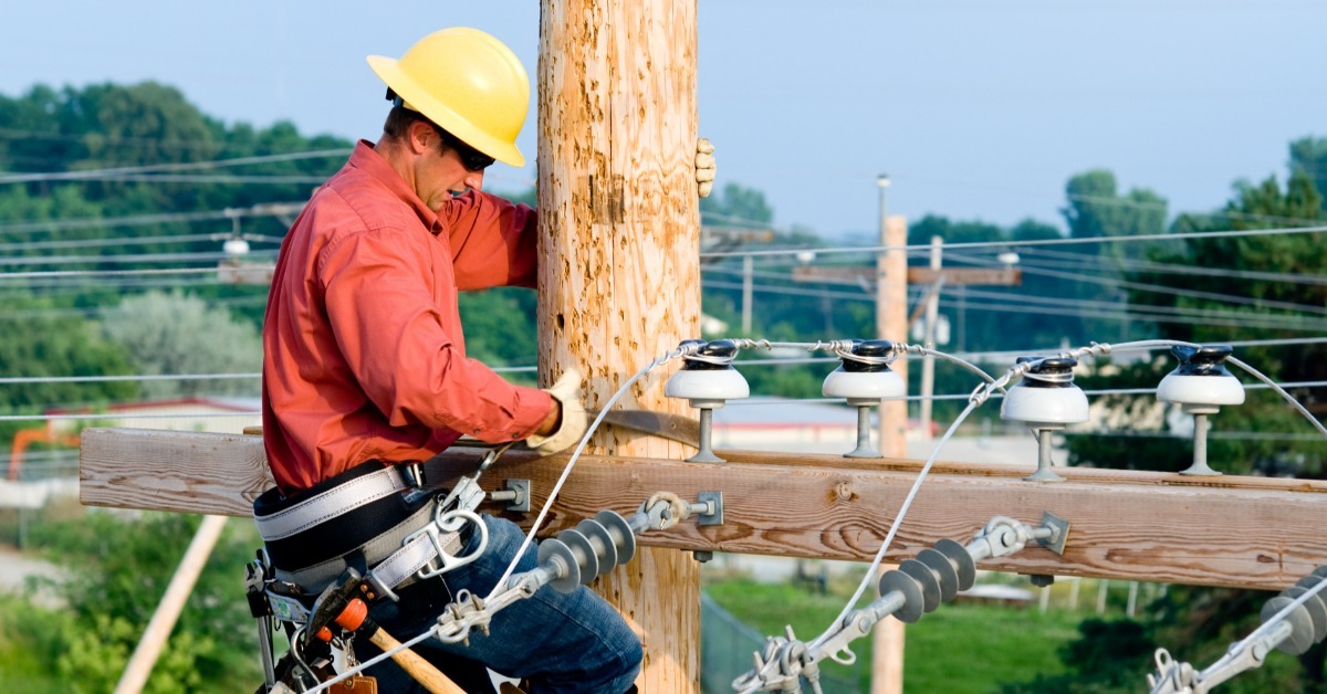 A utility worker wearing a yellow hard hat and safety harness while working on a wooden power pole with electrical wires.