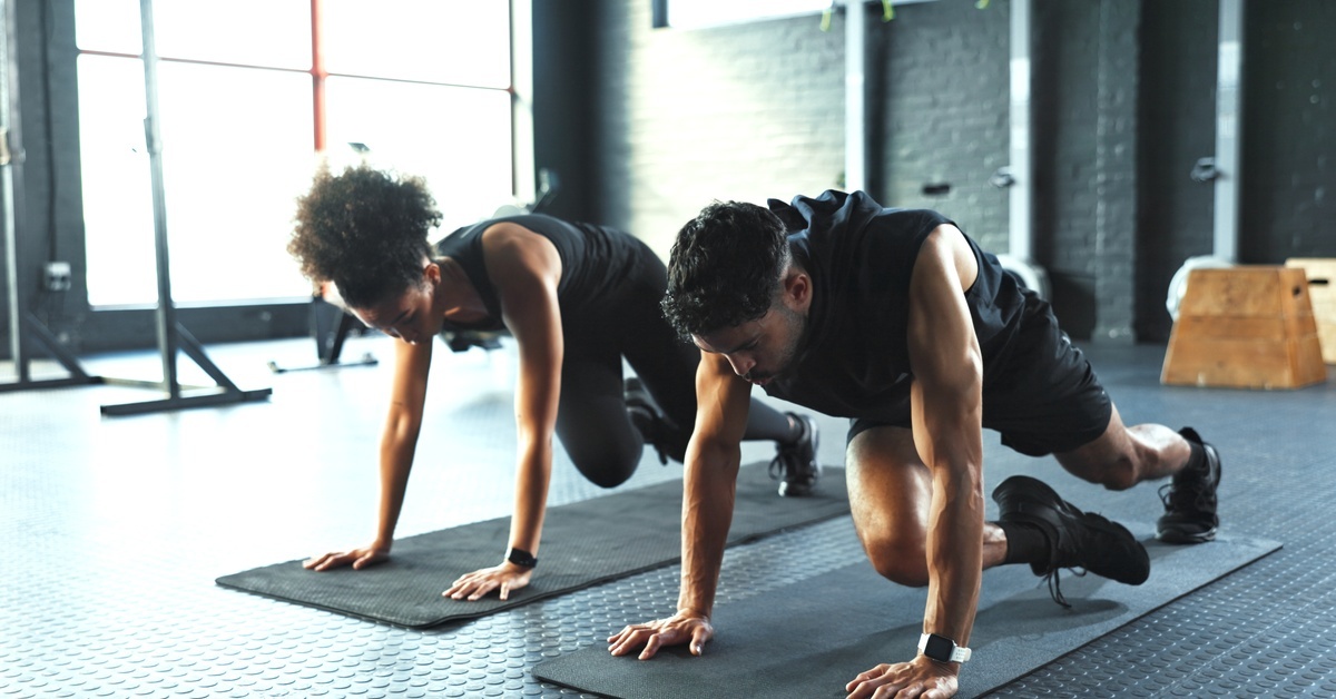 A man and a woman are doing mountain climbers in a gym on black yoga mats. They are wearing black workout gear.