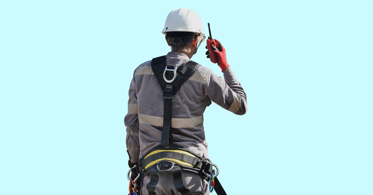 A person wearing a gray jumpsuit with white visibility stripes and a white hard hat is talking into a walkie-talkie.