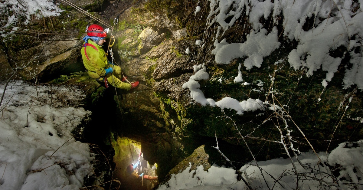 A person in a yellow jumpsuit, connected to several ropes, climbs into a crevice. Their headlamp illuminates the path.