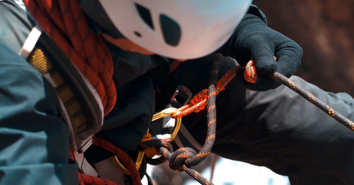 A climber wearing a white helmet while connected to a series of ropes. They are looking at the carabiner hooked to the rope.