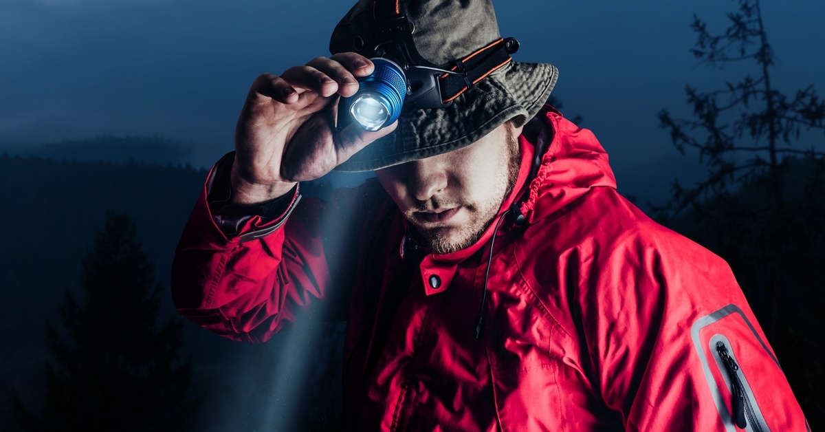 A man in a red jacket and bucket hat wearing a bright headlamp on his head. He uses his right hand to adjust it.