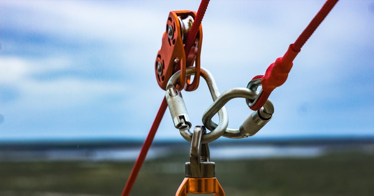 A close-up of three ropes attached to one another with carabiners and a pulley. There is a cloudy sky in the background.
