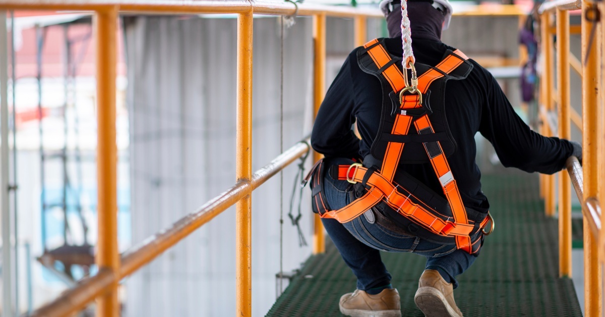 A person wearing a black hoodie and a white hard hat, kneeling on a raised metal catwalk with railing.