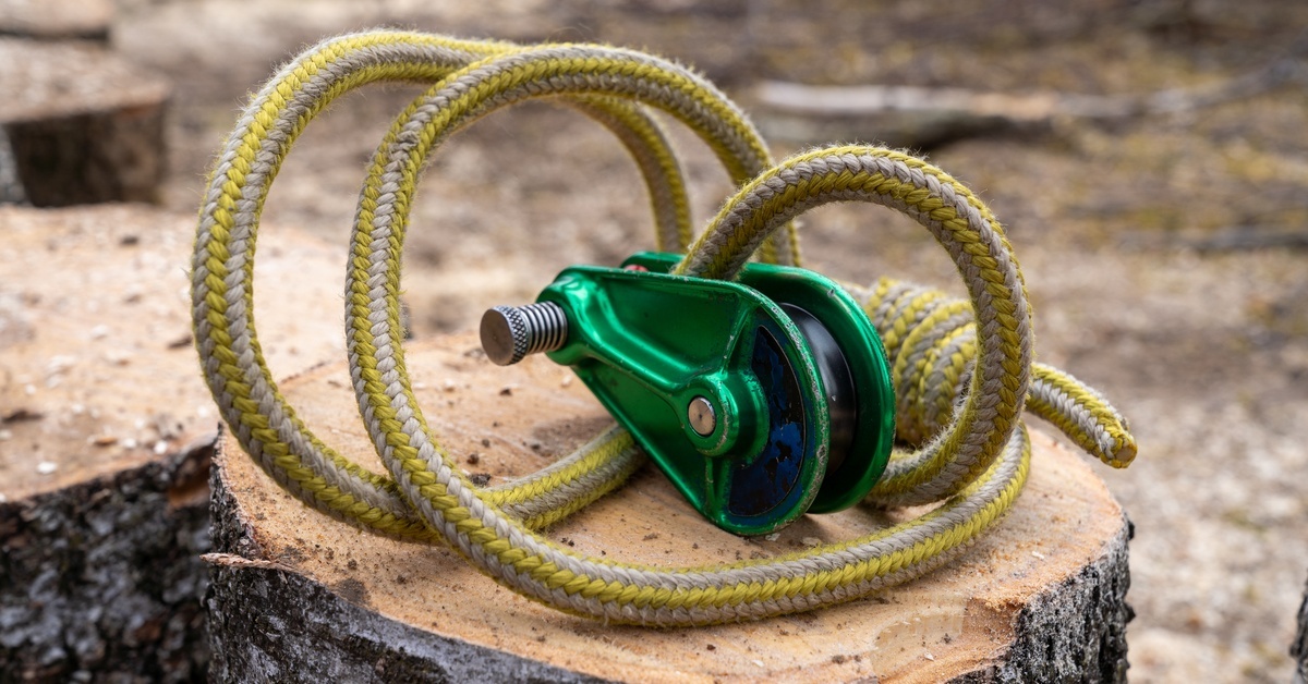 A green arborist rigging block with a coil of yellow and white rope set up on a sawed off section of wood.