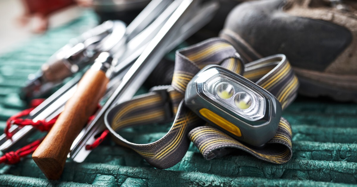A yellow and gray headlamp resting on a green surface next to a blurry pair of boots and some blurry camping gear.