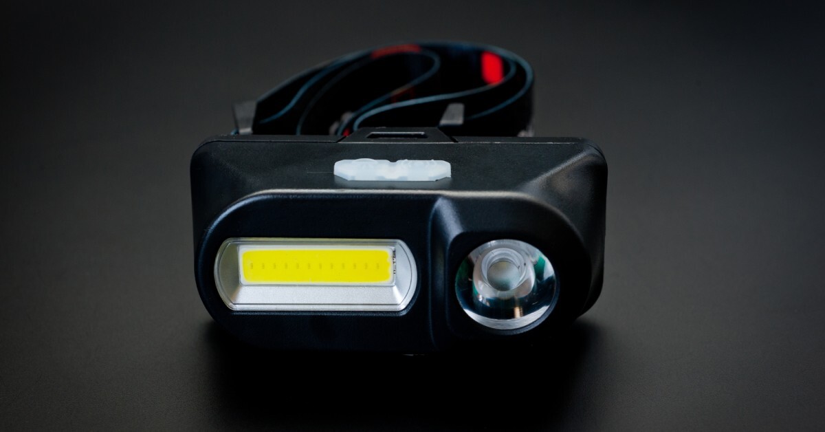 A small black headlamp with a coiled strap resting on a black surface. The headlamp has two light sources.