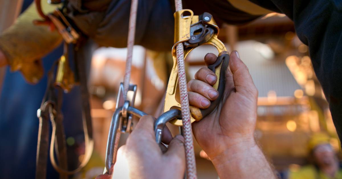 A close-up of a person connecting an orange ascender and a gray carabiner to a rope in front of a person in a harness.