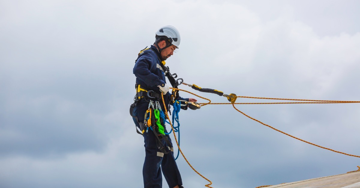 A person in work boots, coveralls, gloves, and a helmet wearing a harness while attached to a rope system.