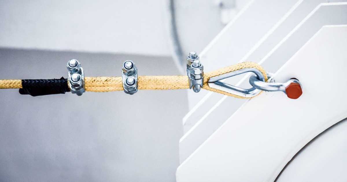 A close-up of a yellow nylon rope with a metal loop and fasteners attached to an anchor point on a metal structure.