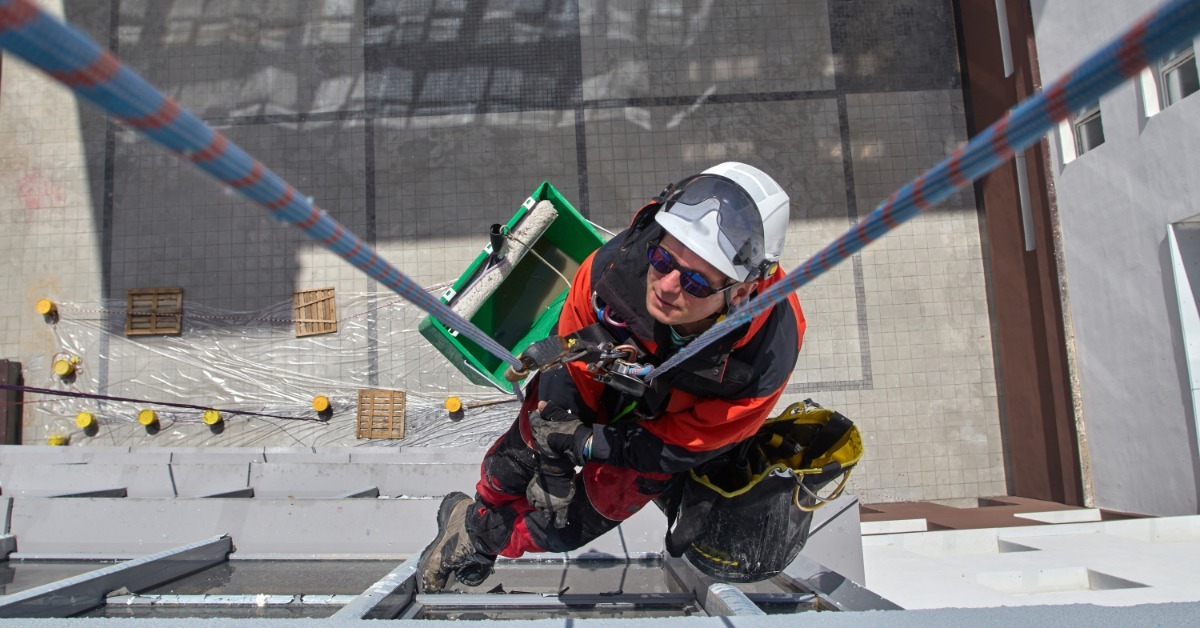 An overhead view of a rope access technician wearing a white helmet and red coveralls while suspended next to a building.
