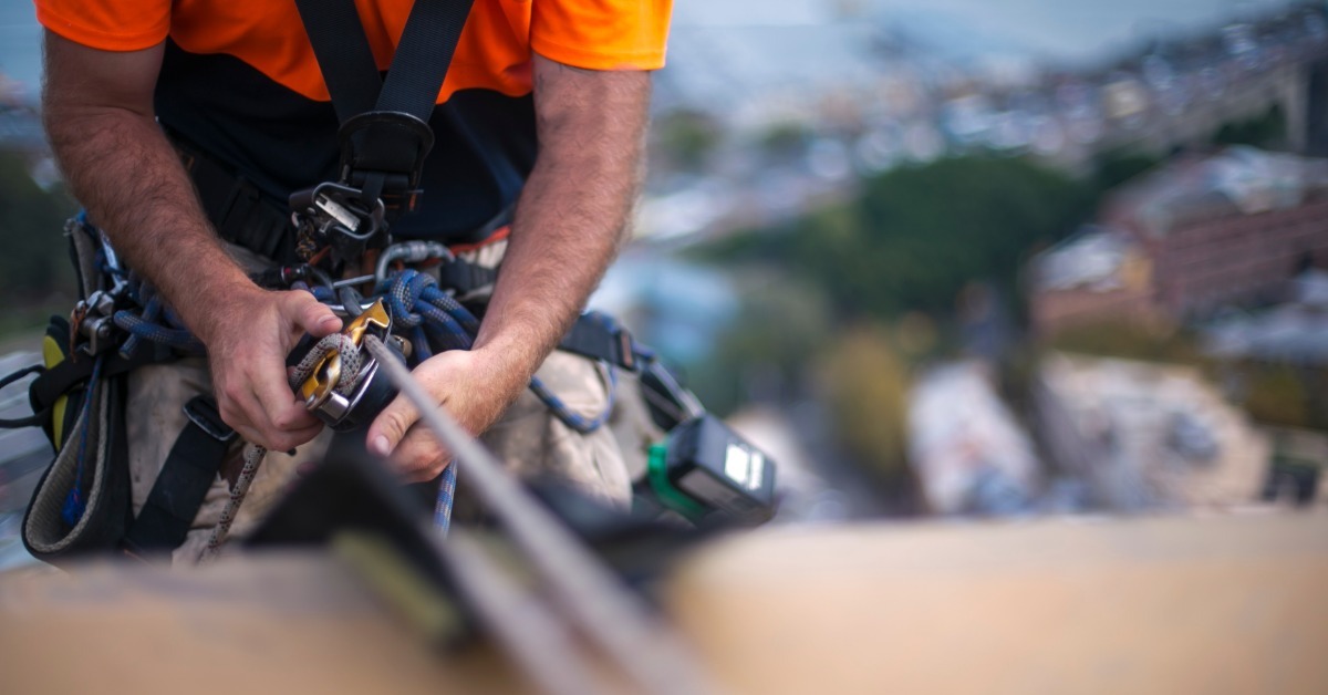 A close-up of a rope access technician wearing a bright orange shirt and a harness while hooked up to a rigging system.