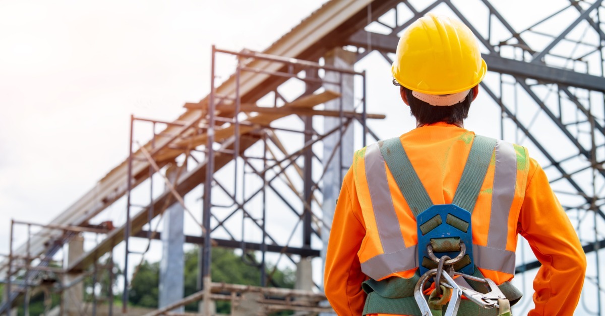 A construction worker wearing a hard hat, a hi-vis shirt, and a harness while looking up at scaffolding on the jobsite.