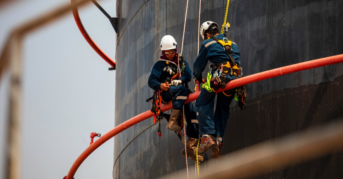 Two workers in safety gear wearing harnesses with attached ropes while sitting on a metal bar on the outside of a large structure.