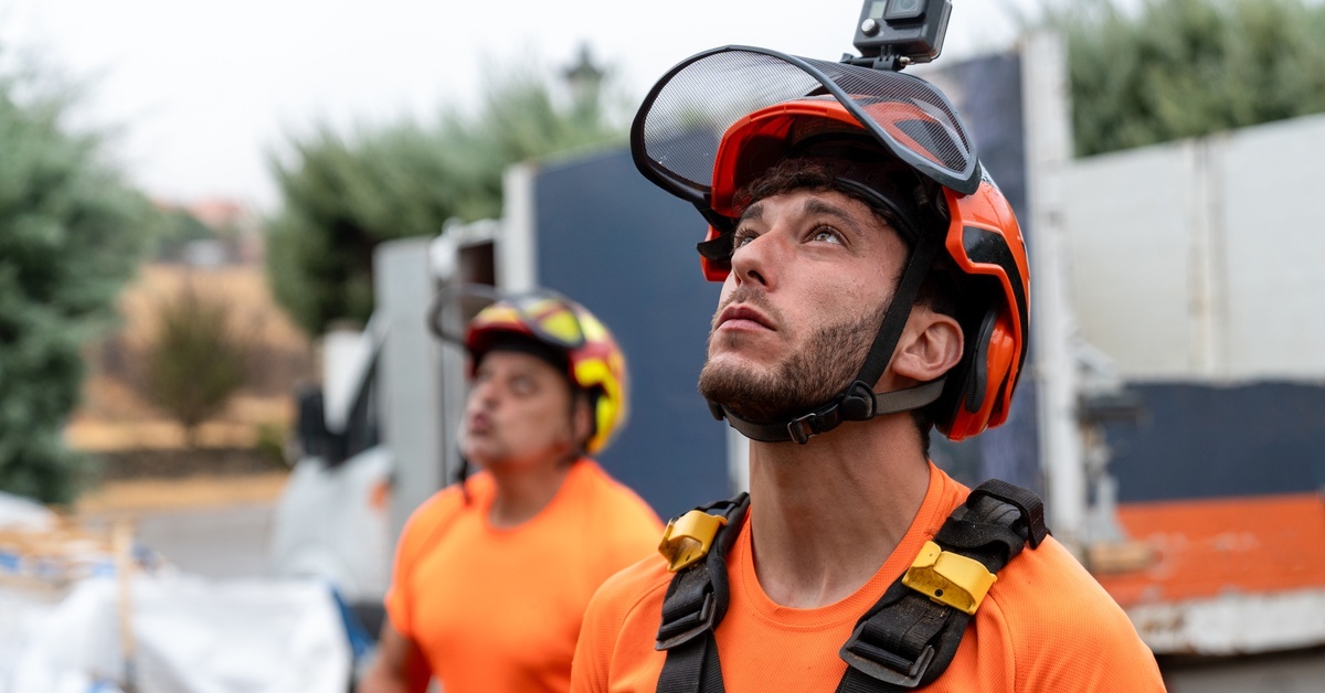 An arborist looking up. He's wearing a hard hat with built-in eye protection, a harness, and an orange T-shirt.